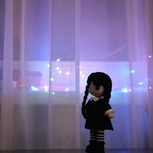 May include: A crocheted doll with black hair, a white collar, and a black dress stands in front of a sheer curtain with colorful lights behind it.