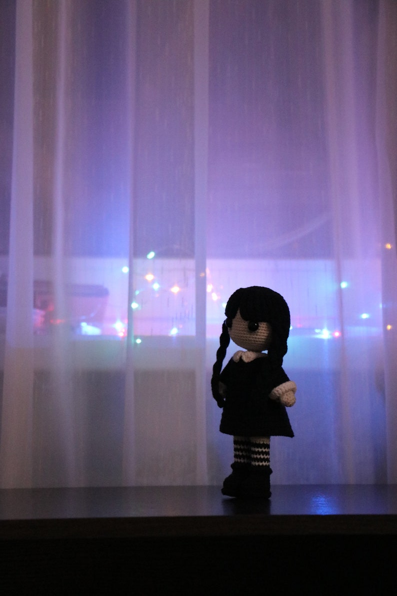 May include: A crocheted doll with black hair, a white collar, and a black dress stands in front of a sheer curtain with colorful lights behind it.