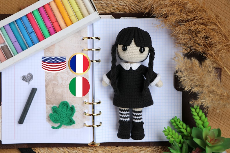May include: A black crocheted doll with black hair, white collar, and black and white striped stockings. The doll is sitting on a notebook with a grid pattern. There are flags of the United States, France, and Italy on the notebook.