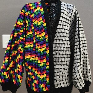 Peut inclure: Cardigan en crochet fait main avec un design unique. Un côté présente un motif arc-en-ciel vibrant, tandis que l'autre côté est un damier en niveaux de gris. Le cardigan a une bordure et des manches noires, et est présenté sur un mannequin.