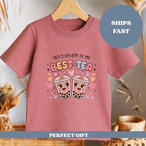 May include: A dusty rose-colored t-shirt featuring a graphic of two smiling bubble tea cups with the text "YOU'LL ALWAYS BE MY BEST-TEA". The design includes hearts and stars. The shirt is hanging on a wooden hanger.