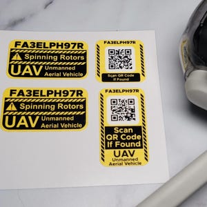 May include: Four yellow and black warning stickers with the text "FA3ELPH97R", "Spinning Rotors", "UAV Unmanned Aerial Vehicle", and "Scan QR Code If Found". Two stickers have a QR code.