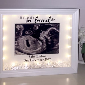 May include: White picture frame with a black and white ultrasound image inside. The frame has a white background with the text "So little so loved" and a heart symbol. The text "Baby Barlow Due December 2022" is also included. The frame is decorated with clear rhinestones and has a string of white lights around the edge.