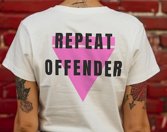 May include: A white t-shirt with the words "REPEAT OFFENDER" in black text above a pink triangle. The shirt is worn by a person with tattoos on their arms, against a red brick wall background.