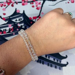 Puede incluir: Dos pulseras de cuentas transparentes se usan en una muñeca, con una pulsera plateada visible en la muñeca inferior. Las cuentas tienen una ligera cualidad iridiscente. Un anillo plateado con una piedra turquesa está en un dedo.