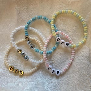 May include: Four beaded bracelets in pastel colors: cream, light blue, pink, and yellow. Each bracelet has the letters "CTR" spelled out with gold or white letter beads. The bracelets are made of small, round beads.
