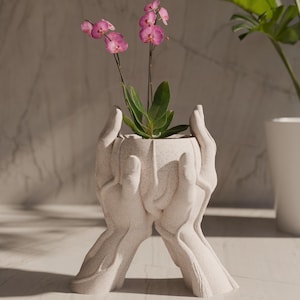 Könnte beinhalten: Ein weißer Keramik-Pflanztopf in Form von zwei Händen, die eine kleine Pflanze mit rosa Blüten halten. Der Topf steht auf einer weißen Oberfläche.
