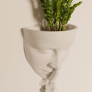 Peut inclure: Plante verte poussant dans un pot en céramique blanche en forme de visage. Le visage a les yeux fermés et le doigt sur les lèvres, comme pour dire "chut".