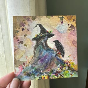 May include: A square watercolor painting depicting a figure wearing a pointed hat adorned with small flowers, accompanied by a black raven perched on their shoulder. The scene features a colorful, textured background with autumn-toned leaves.
