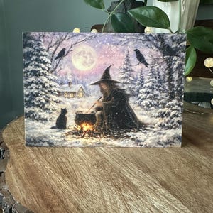 May include: A rectangular art print depicting a witch stirring a cauldron in a snowy forest. A black cat sits nearby. The scene includes a full moon, two crows, and a small house. The color palette is dominated by whites, blues, and browns.