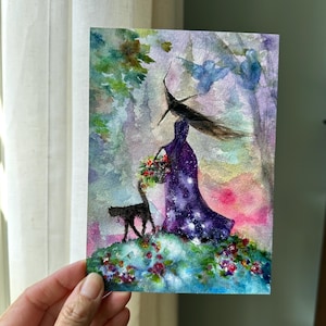 May include: A watercolor painting on paper depicting a figure in a long, dark purple gown with white star-like speckles, wearing a pointed hat. A black cat stands nearby on a grassy patch filled with colorful flowers against a soft, hazy background.