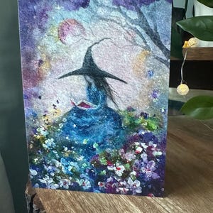 May include: A watercolor print of a witch reading a book, set in a whimsical garden. The witch wears a black hat and blue dress, surrounded by colorful flowers and a soft, starry background. A pink moon and tree branches are visible.