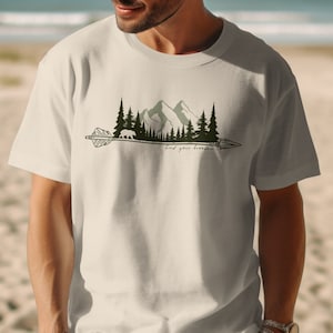 May include: A white t-shirt with a green mountain and forest design. The design includes a bear walking through the forest and an arrow with the text "find your freedom".