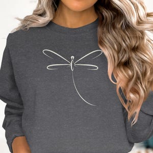 May include: Charcoal grey sweatshirt with a minimalist white dragonfly design. The dragonfly is rendered in simple, elegant lines, creating a clean and modern aesthetic. The sweatshirt is a comfortable, casual style.