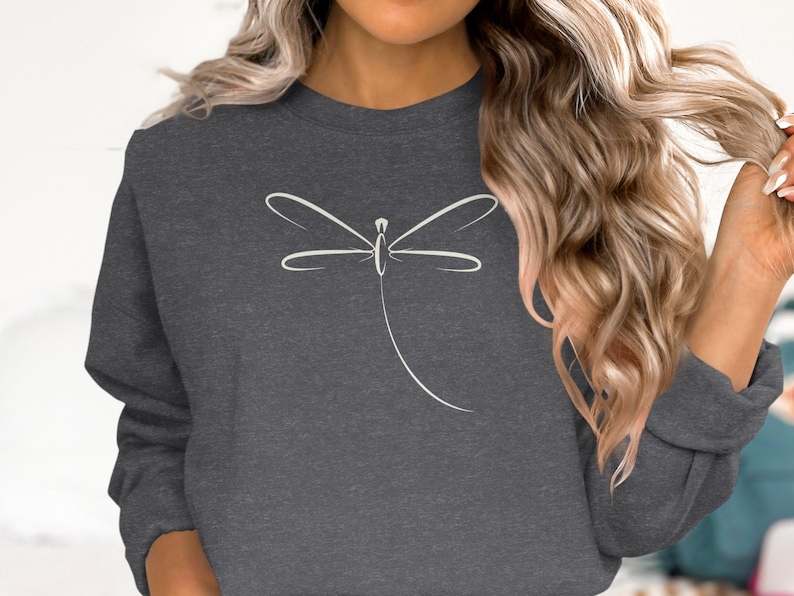 May include: Charcoal grey sweatshirt with a minimalist white dragonfly design. The dragonfly is rendered in simple, elegant lines, creating a clean and modern aesthetic. The sweatshirt is a comfortable, casual style.