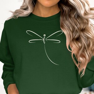 May include: A dark green sweatshirt with a white dragonfly outline. The dragonfly design is minimalist and stylish. The sweatshirt has a round neck and long sleeves, ideal for everyday wear. The colour is a deep forest green.
