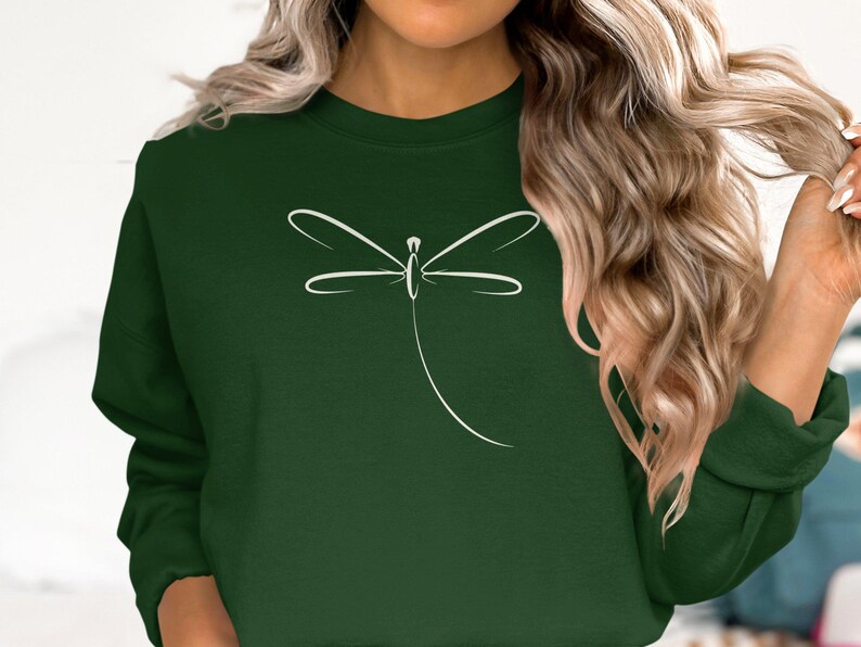 May include: A dark green sweatshirt with a white dragonfly outline. The dragonfly design is minimalist and stylish. The sweatshirt has a round neck and long sleeves, ideal for everyday wear. The colour is a deep forest green.
