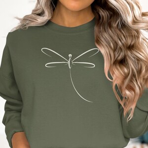 May include: An olive green sweatshirt with a white dragonfly design. The dragonfly is drawn with simple lines, giving a clean, modern look. The sweatshirt has long sleeves and a crew neck. The colour is a muted, earthy tone.