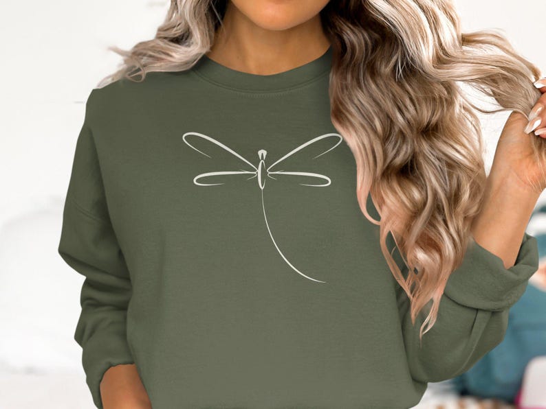 May include: An olive green sweatshirt with a white dragonfly design. The dragonfly is drawn with simple lines, giving a clean, modern look. The sweatshirt has long sleeves and a crew neck. The colour is a muted, earthy tone.