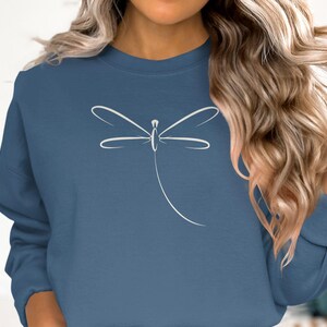 May include: A blue sweatshirt with a white dragonfly design. The dragonfly is illustrated with fine lines, giving a minimalist and elegant look. The sweatshirt is a solid colour, offering a clean background for the graphic.