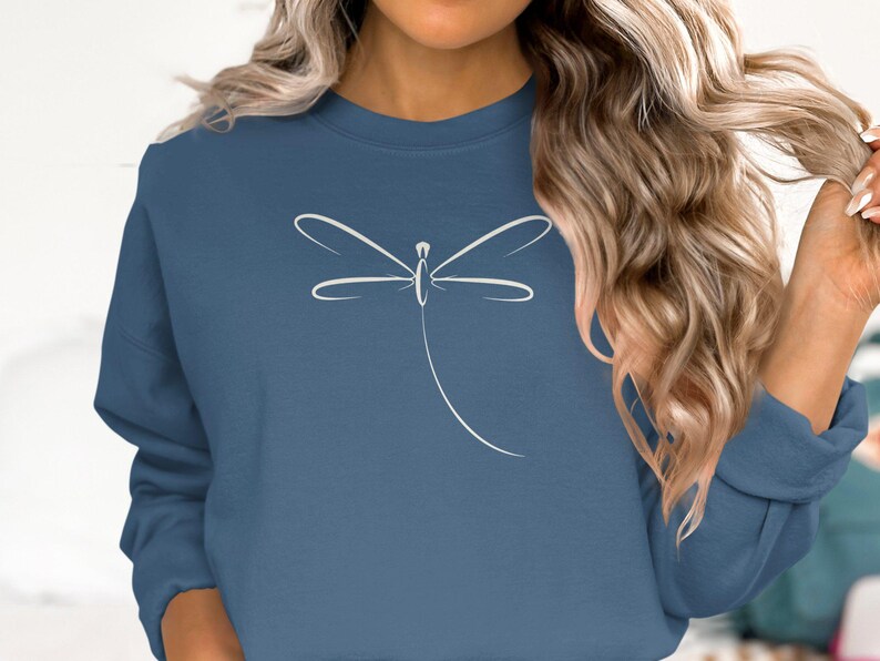 May include: A blue sweatshirt with a white dragonfly design. The dragonfly is illustrated with fine lines, giving a minimalist and elegant look. The sweatshirt is a solid colour, offering a clean background for the graphic.