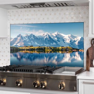May include: A stainless steel stovetop with a black grate and gold knobs. The stovetop is set against a backdrop of a mountain range reflected in a still lake.