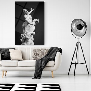 Pu&ograve; includere: Una fotografia in bianco e nero di una scultura che rappresenta tre figure in una posa drammatica. La scultura &egrave; esposta su una parete bianca in un soggiorno moderno. Un divano bianco con cuscini neri e beige &egrave; in primo piano, e una lampada da terra a treppiede nera con un riflettore argentato &egrave; sullo sfondo.