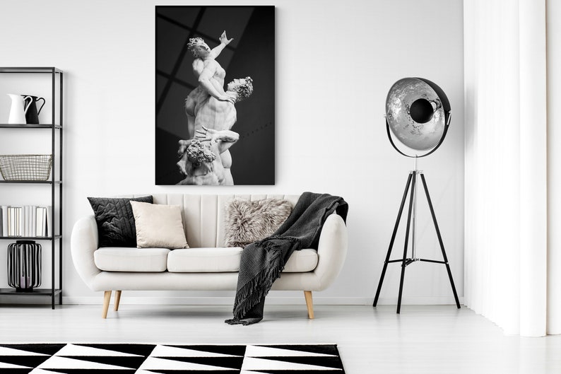Pu&ograve; includere: Una fotografia in bianco e nero di una scultura che rappresenta tre figure in una posa drammatica. La scultura &egrave; esposta su una parete bianca in un soggiorno moderno. Un divano bianco con cuscini neri e beige &egrave; in primo piano, e una lampada da terra a treppiede nera con un riflettore argentato &egrave; sullo sfondo.