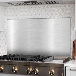 May include: Stainless steel range hood and backsplash. The backsplash is a large, brushed metal panel. The range has multiple burners and gold-colored knobs. The background features white and gold geometric tile.