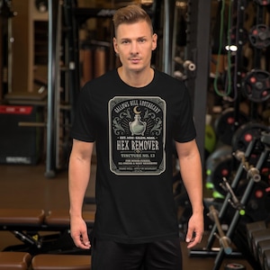 May include: A black short-sleeved t-shirt featuring a graphic label design that reads Gallows Hill Apothecary, Est. 1692, Salem, Mass., Hex Remover, Tincture No. 13, For Minor Curses, Ill Omens & Nosy Neighbors, Shake Well, Apply By Moonlight.