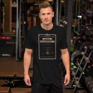 May include: A black short-sleeve t-shirt featuring a graphic print of an annual performance review document. The text reads "ANNUAL PERFORMANCE REVIEW" and "EXCEEDS EXPECTATIONS" in a decorative frame with a gold-colored seal.