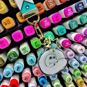 May include: A collection of colorful art markers with a variety of shades, including pink, green, and blue. A white lanyard with a teal and gold accent is attached to a gold keychain with a cartoon character charm.