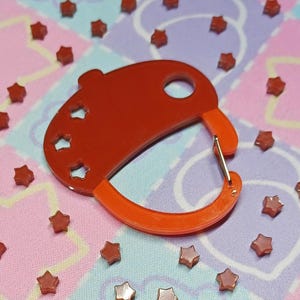 Può includere: Un moschettone rosso con ritagli a forma di stella e una chiusura arancione. Piccoli abbellimenti a forma di stella sono sparsi su uno sfondo pastello a fantasia. Un accessorio funzionale.