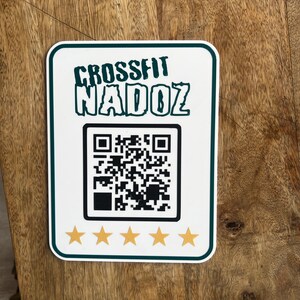Peut inclure: Un panneau blanc avec du texte vert qui dit "CrossFit NADOZ". Un code QR noir et blanc se trouve sous le texte. Quatre étoiles dorées se trouvent en bas du panneau.
