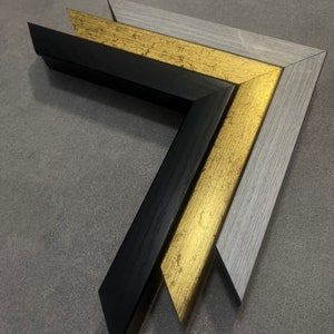 May include: Three picture frames, one black, one gold, and one gray, are stacked on top of each other. The frames are all the same size and shape, and they are all made of wood.