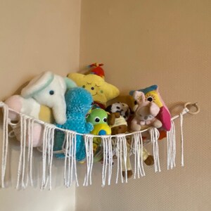 Könnte beinhalten: Ein weißer Makramee-Hängesessel mit Fransen hängt an der Ecke einer Wand. Er ist gefüllt mit verschiedenen Stofftieren, darunter ein blauer Oktopus, ein gelber Stern, ein rosa Elefant und ein brauner Bär.