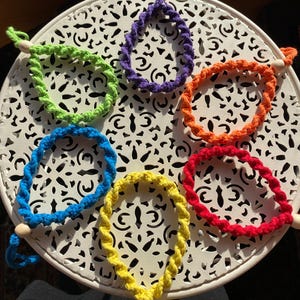 Könnte beinhalten: Sechs bunte gehäkelte Armbänder mit Holzperlen. Die Armbänder sind in einem Kreis auf einem weißen Tisch mit einem dekorativen Muster angeordnet.