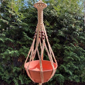 Könnte beinhalten: Ein hängender Makramee-Pflanzenhalter mit einem terrakottafarbenen Topf. Das Makramee besteht aus naturfarbenem Seil mit dunkelbraunen Perlen. Der Pflanzenhalter hängt, mit grünem Laub im Hintergrund.