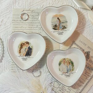 Puede incluir: Tres platos de cerámica blanca en forma de corazón con ilustraciones personalizadas de parejas. Cada plato muestra un retrato único, los nombres de la pareja, la palabra Engaged y una fecha. Los platos tienen un acabado liso y bordes plateados.