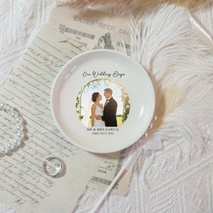 May include: A white ceramic plate featuring a wedding photo of a couple under an archway. The plate is inscribed with "Our Wedding Days", the couple's names, and the date "20rd July 2026". The plate is surrounded by decorative elements.