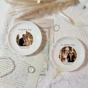 May include: Two white ceramic plates with wedding photos and text. Each plate features a couple's photo, the phrase "Our Wedding Day", and names with a date. The plates are on a textured surface with decorative elements.