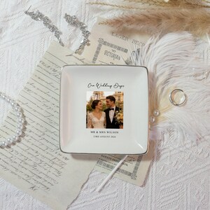 May include: A square, white ceramic ring dish with a gold trim. The dish features a photograph of a couple and the text "One Wedding Day", "MR & MRS WILSON", and "23RD AUGUST 2026". The dish is surrounded by vintage ephemera and jewelry.