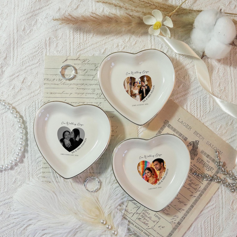 May include: Three heart-shaped ceramic dishes with a silver trim. Each dish features a wedding photo and the text "Our Wedding Days" with a date and names. The dishes are on a textured white surface with decorative elements.