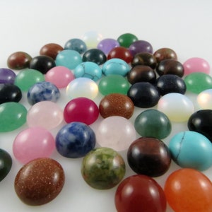 May include: Assortment of colorful cabochon gemstones in various shapes and sizes. The gemstones are arranged in a pile on a white background.