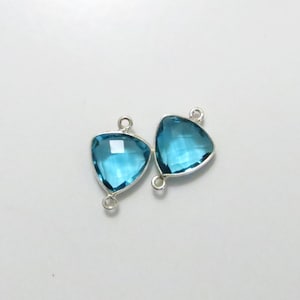 May include: Two silver triangle-shaped connector charms with a blue topaz gemstone in the center. The charms have two loops on the side for attaching to jewelry.