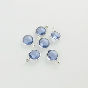 May include: Six round, blue gemstone pendants with silver-colored metal settings and small loops. The pendants are arranged on a white surface, showcasing their faceted surfaces and reflective qualities. The gemstones have a light blue hue.