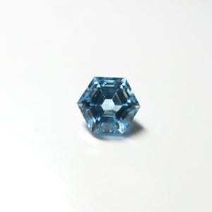 May include: A faceted, hexagonal gemstone with a light blue hue. The gem has a symmetrical design with multiple facets that reflect light. The gemstone is set against a white background.