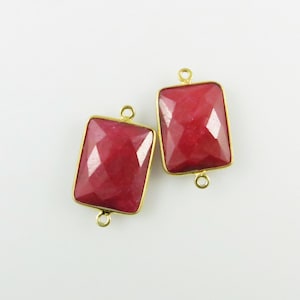 Może przedstawiać: Dwa złote prostokątne charmsy łączące z czerwonymi wstawkami z kamieni szlachetnych. Charmsy mają po dwa pierścienie skokowe po każdej stronie.