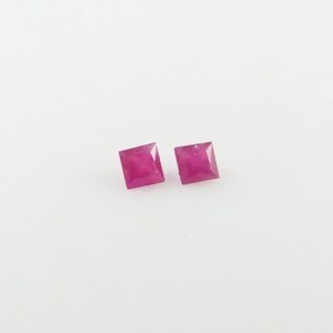Op de afbeelding: Twee vierkante roze edelstenen, waarschijnlijk robijnen, tegen een witte achtergrond.