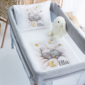 Può includere: Un set di biancheria da letto per culla bianco con bordo grigio. La biancheria da letto presenta un'illustrazione ad acquerello di un orso che dorme su una luna con le stelle. La biancheria da letto include anche un cuscino con lo stesso design e il nome "Ella" stampato su di esso. È incluso anche un coniglietto di peluche bianco.